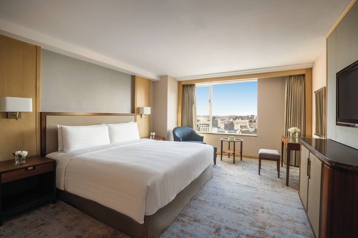 A hotel room with a large bed, a desk, a chair, a TV, and a window with a city view.