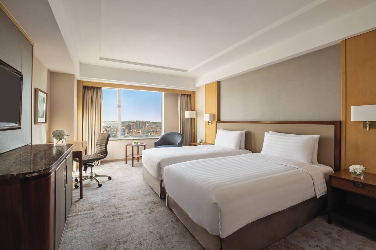 A hotel room with two beds, a desk, a chair, a TV, and a view of the city.