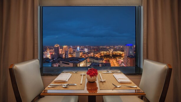 A dining table with a vase of red flowers, set with cutlery and napkins, facing a large window with a cityscape view at dusk. - Shangri-La Changchun (Changchun)