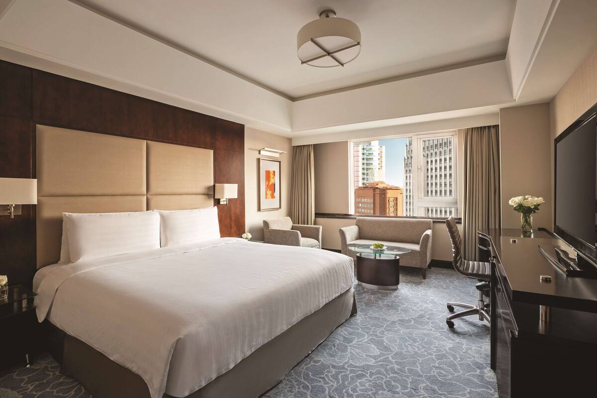 A hotel room with a large bed, a desk, a chair, a small table, and a TV.