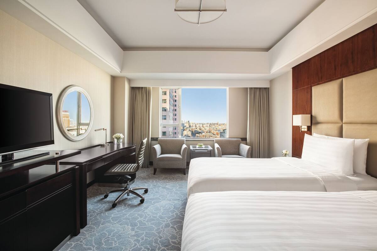 A hotel room with a large bed, a desk with a TV, a chair, a small table, and a view of the city through the window.
