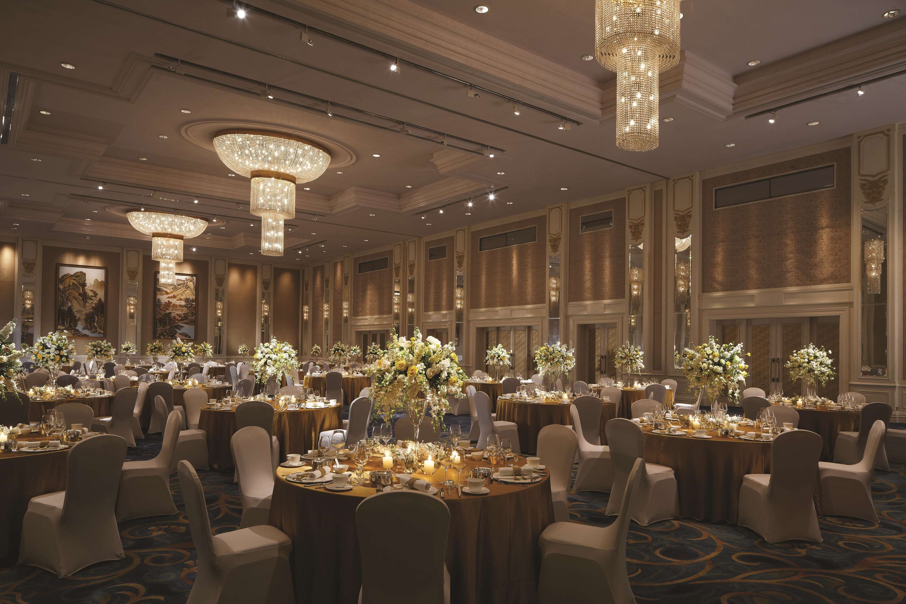 A banquet hall with round tables, chandeliers, and floral centerpieces.
