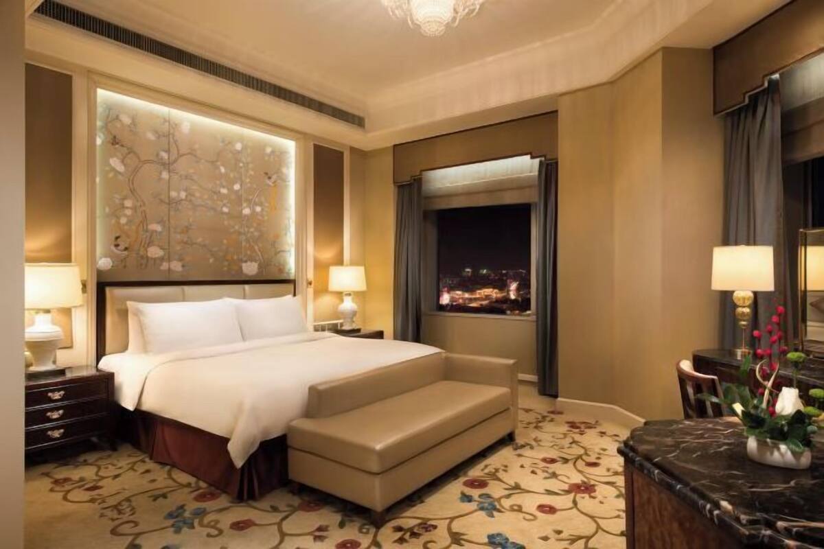 A hotel room with a bed, bedside tables, a sofa, a marble countertop, and a view of the city at night.