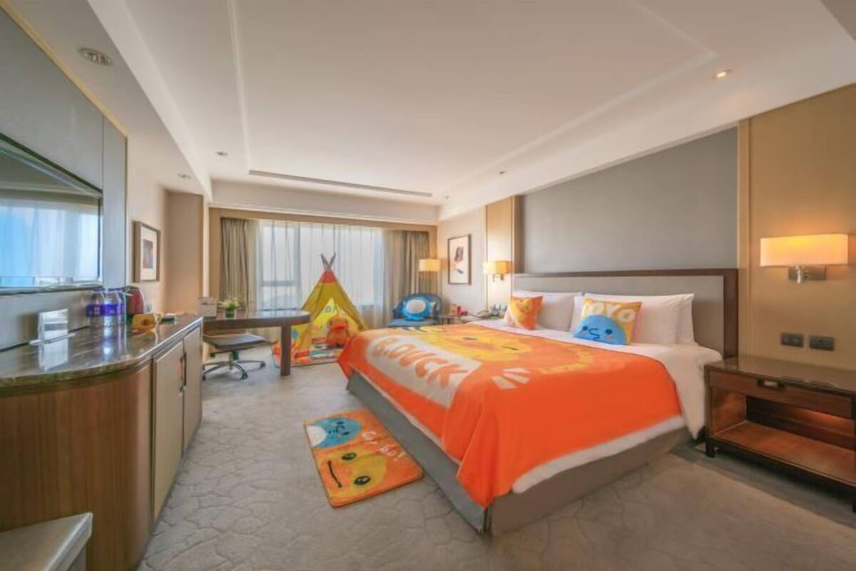 A hotel room with a bed, a desk, a chair, a small table, a lamp, a window with curtains, and a wall-mounted TV.
