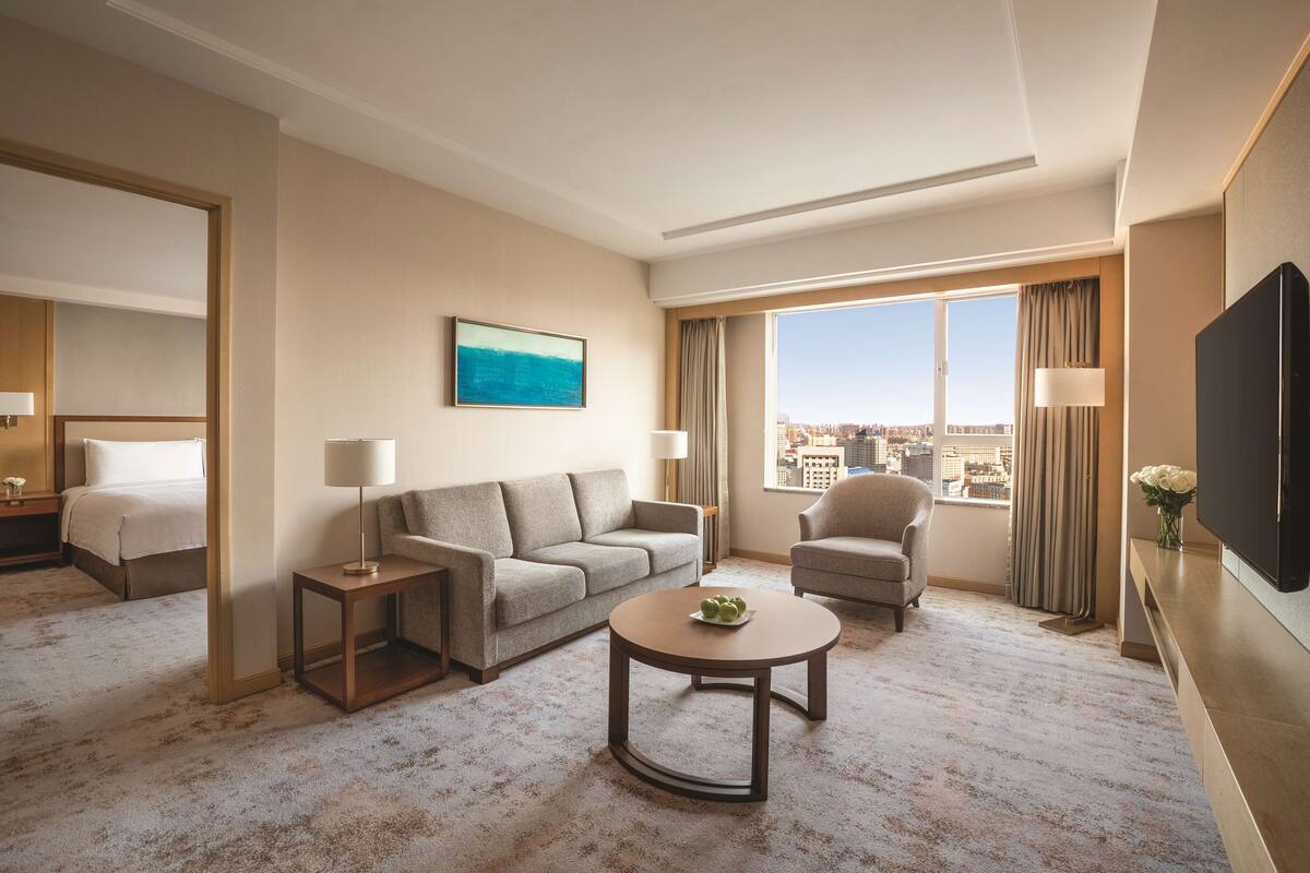 A hotel room with a bed, a sofa, a chair, a coffee table, a TV, and a large window with a city view.