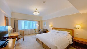 Habitación de hotel con cama, escritorio con televisor, silla, lámpara y ventana con cortinas.