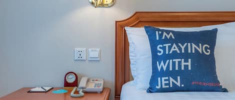 Une chambre d’hôtel avec une table de chevet en bois, une lampe, une horloge, un téléphone et un lit avec un oreiller sur lequel on peut lire : « JE LOGE CHEZ JEN. »