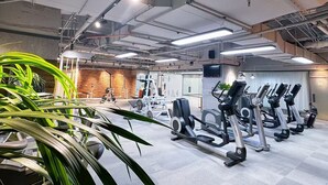 Un gimnasio con diverso equipo de ejercicio, incluyendo cintas de correr y máquinas de pesas, y una pared de ladrillo al fondo.