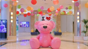 Un vestíbulo amplio con una gran escultura de un oso de peluche rosa, coloridos farolillos de papel colgantes, una pantalla de televisión y una planta en maceta.