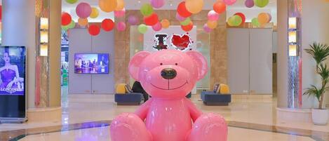 A spacious lobby with a large pink teddy bear sculpture, colorful hanging paper lanterns, a television screen, and a potted plant.