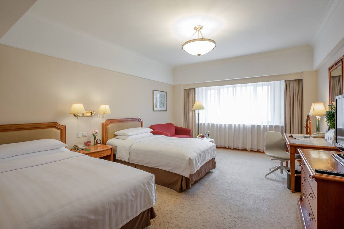A hotel room with two beds, a desk, a chair, a television, and a large window with curtains.