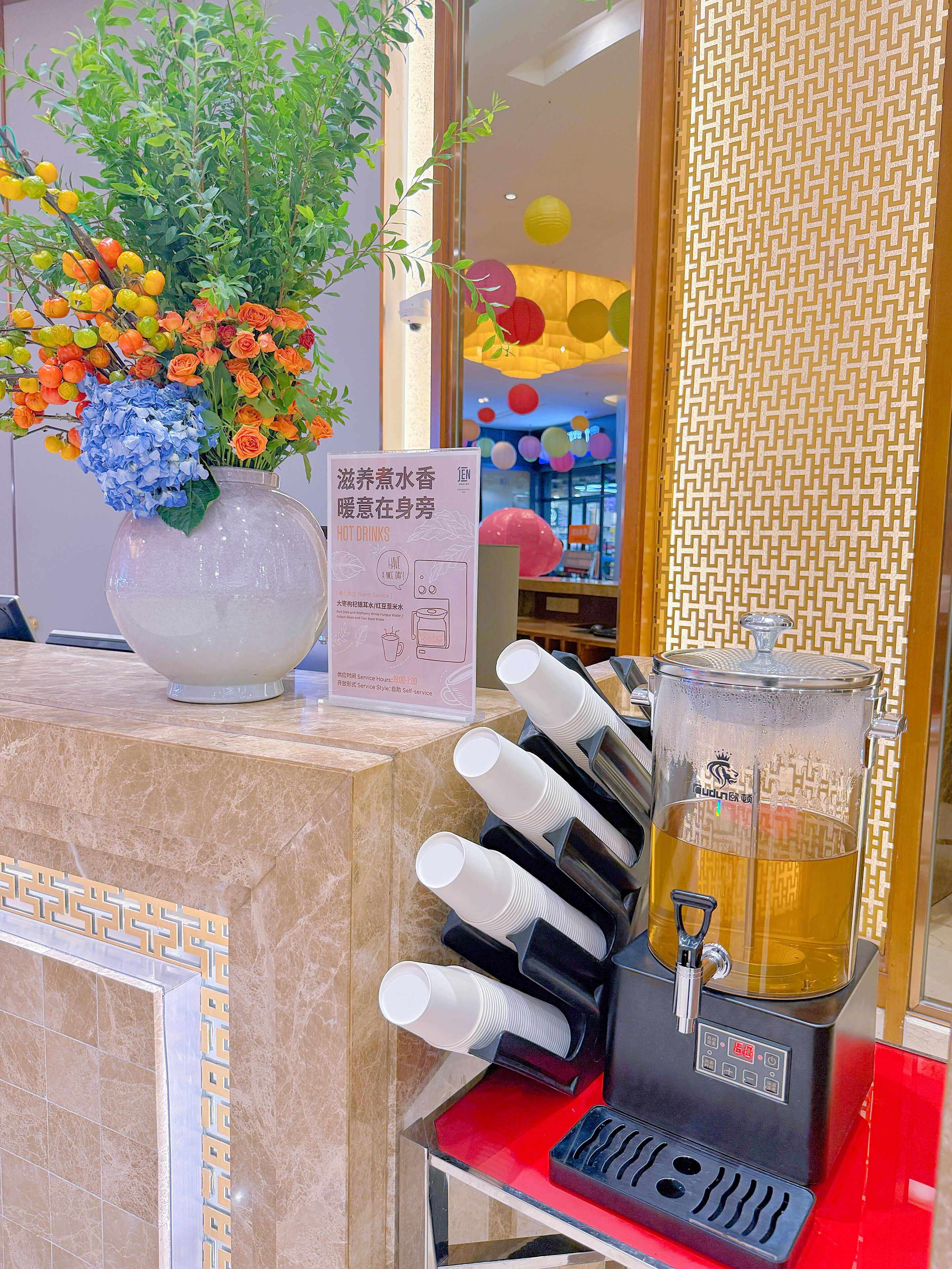 A reception area with a floral arrangement, a beverage dispenser, and a sign indicating hot drinks service hours.