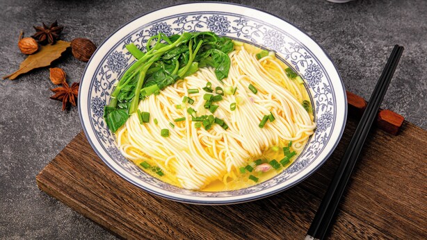 A bowl of noodles with green vegetables and herbs, placed on a wooden board. - JEN Shenyang by Shangri-La (Shenyang)