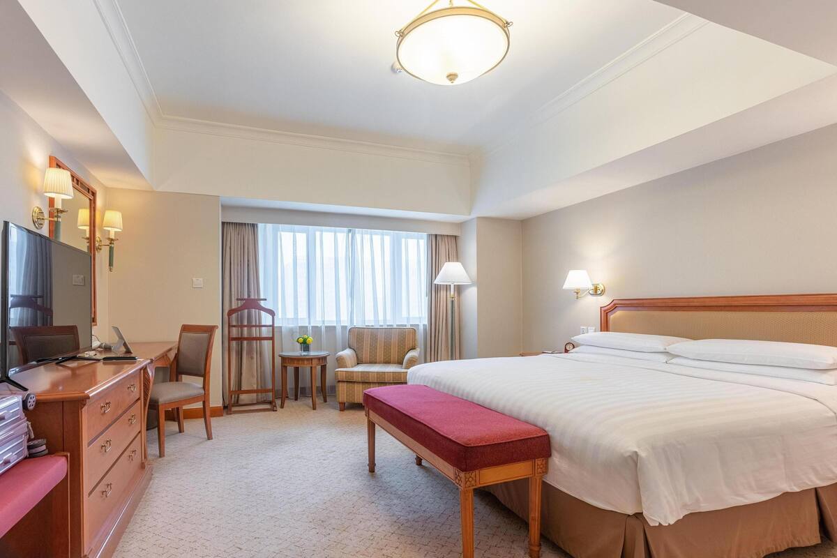 A hotel room with a large bed, a desk with a computer, a chair, a small table with a lamp, and a mirror.