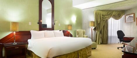 Quarto de hotel com uma cama grande, uma escrivaninha, uma cadeira, um abajur e um quadro na parede.