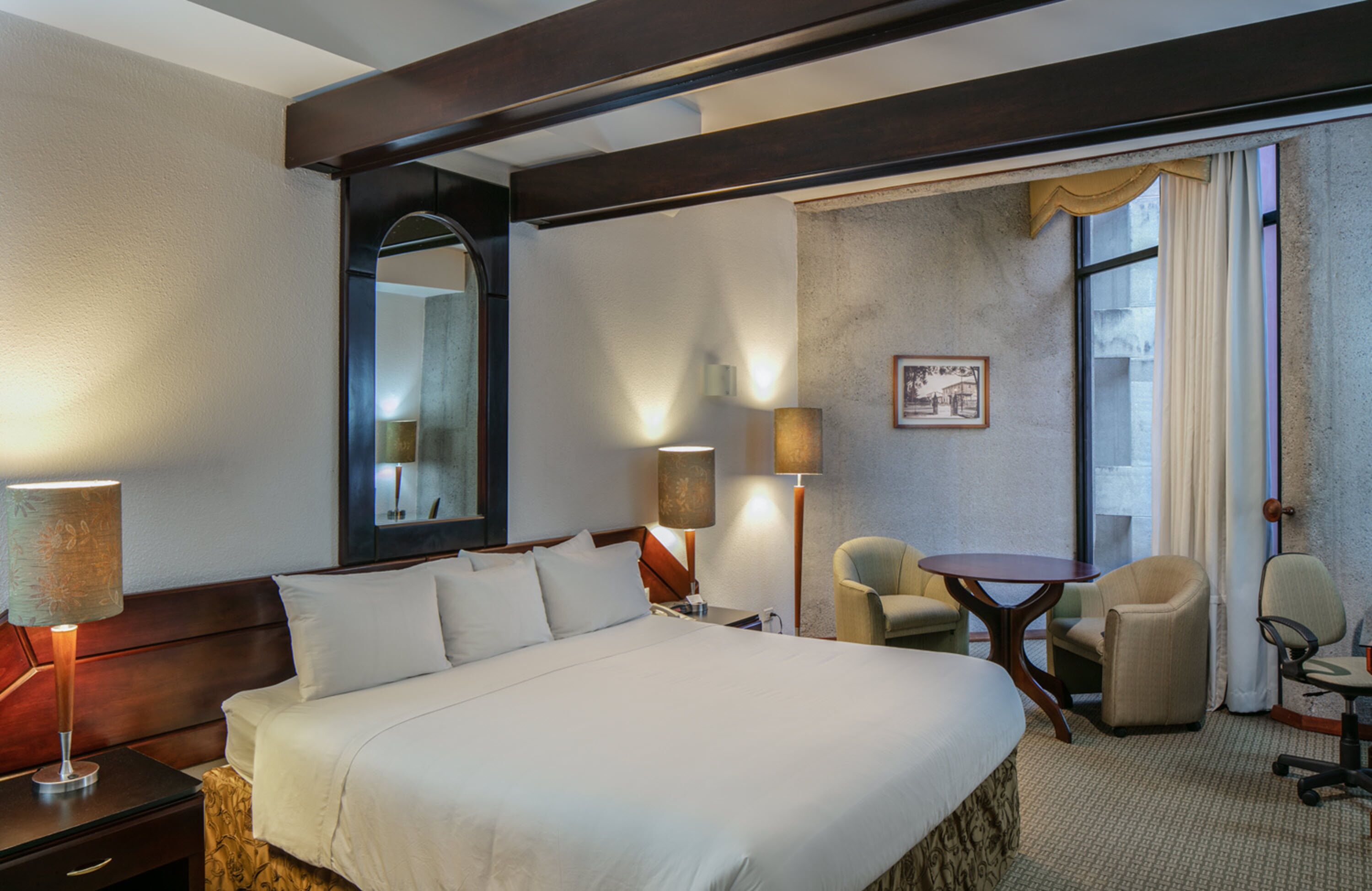 A hotel room with a large bed, two chairs, a small table, and a mirror.
