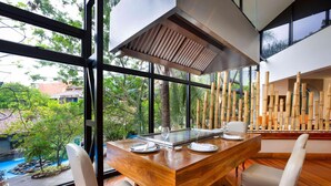 A dining area with a wooden table, white chairs, and a view of a garden and pool through large windows.