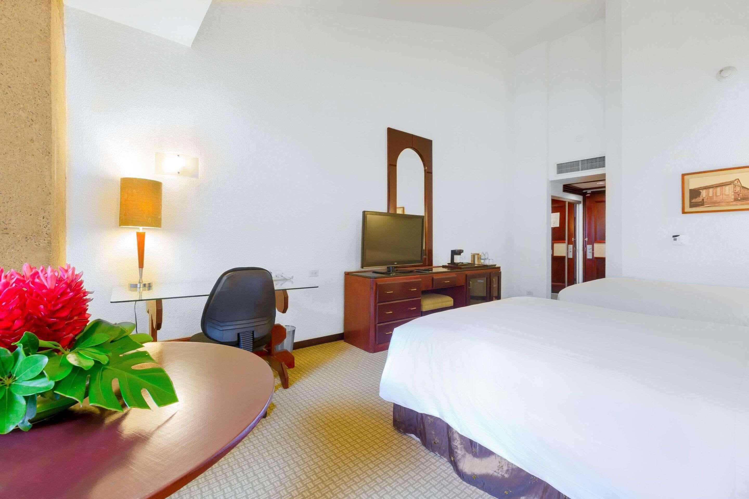 A hotel room with a bed, a desk with a computer, a chair, a TV, a mirror, and a painting on the wall.