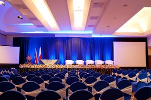 A conference room with rows of chairs, a stage with a blue curtain, and a projector screen.