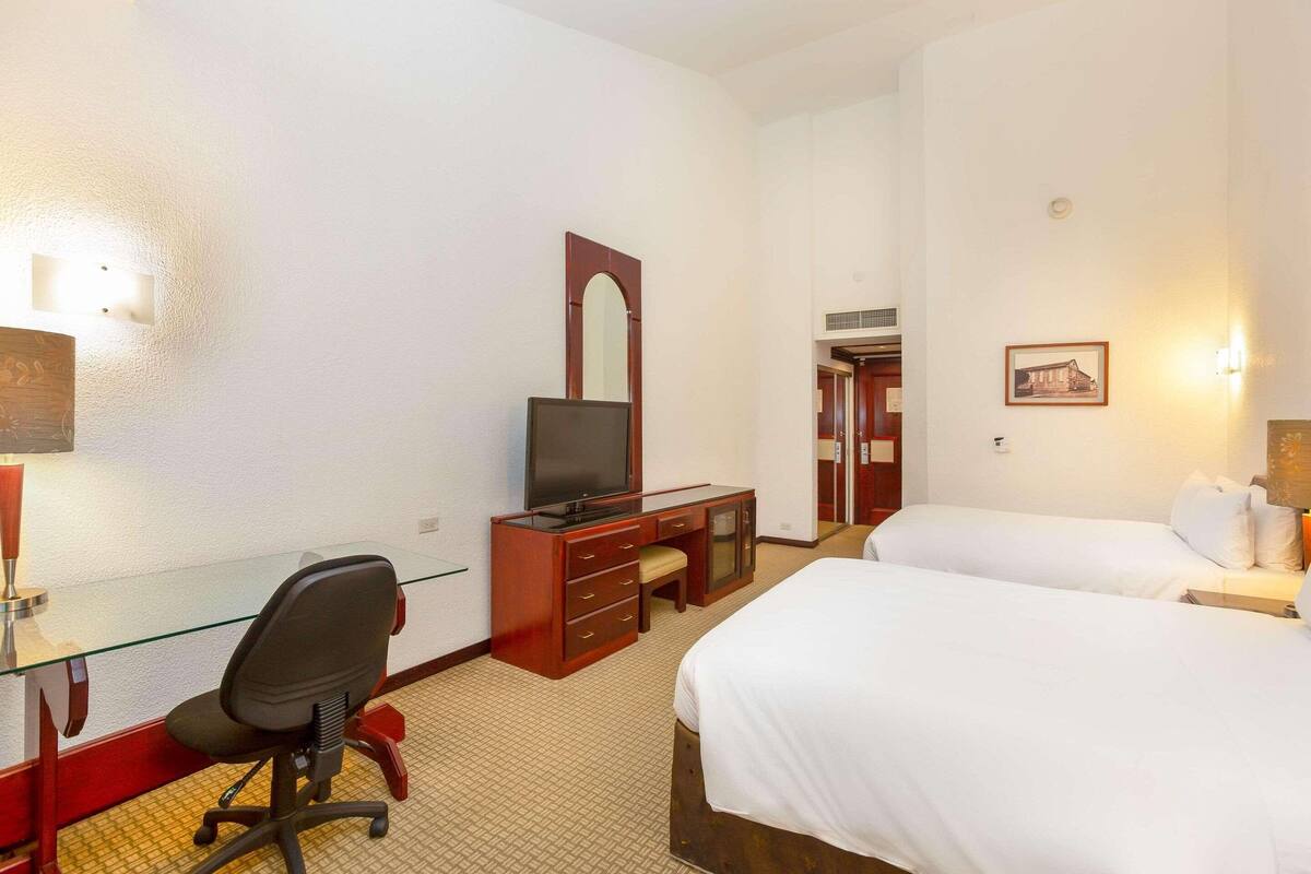 A hotel room with a glass desk, a television, two beds, and a mirror.