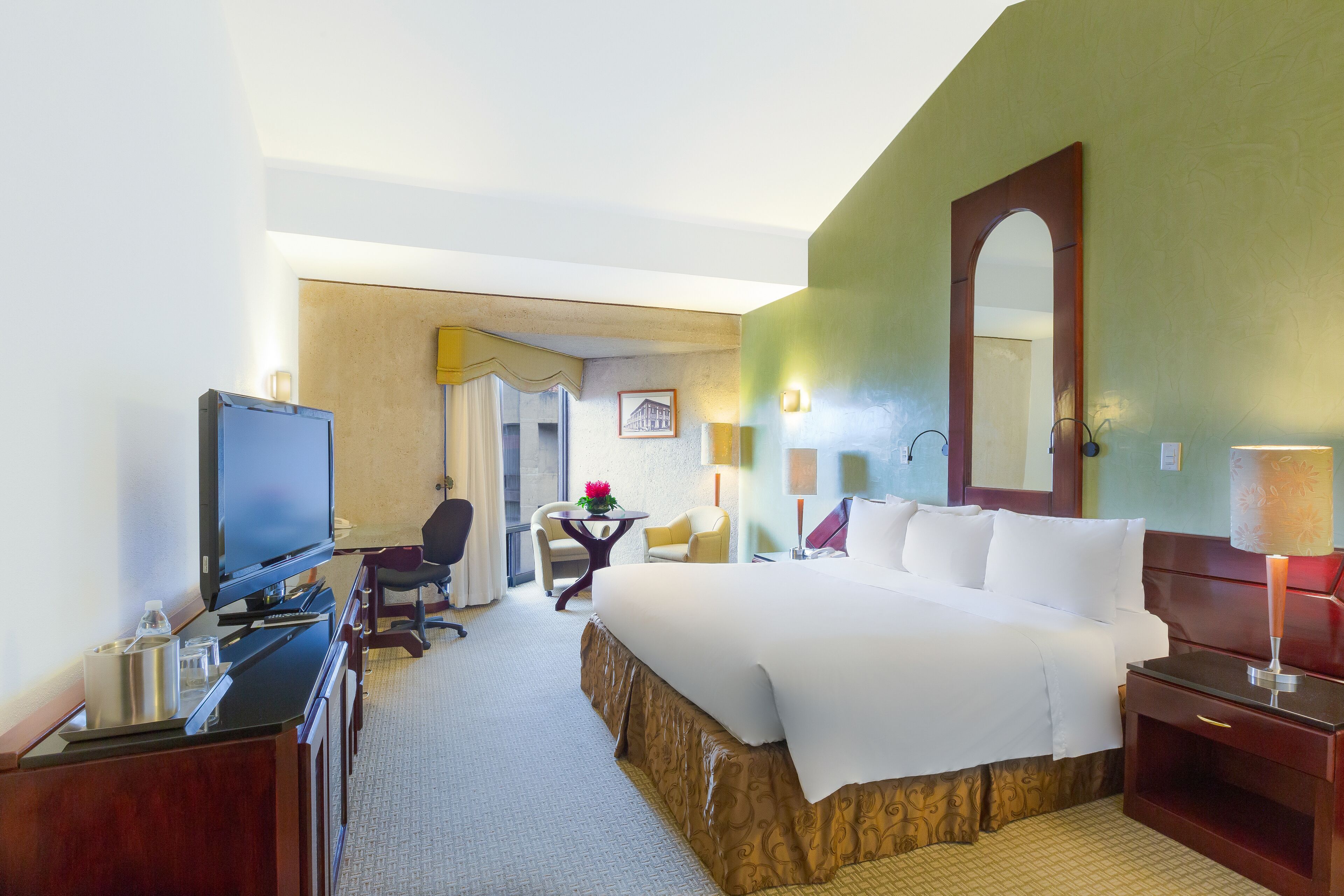A hotel room with a bed, a desk with a television, a chair, a small table with a flower, and a lamp.