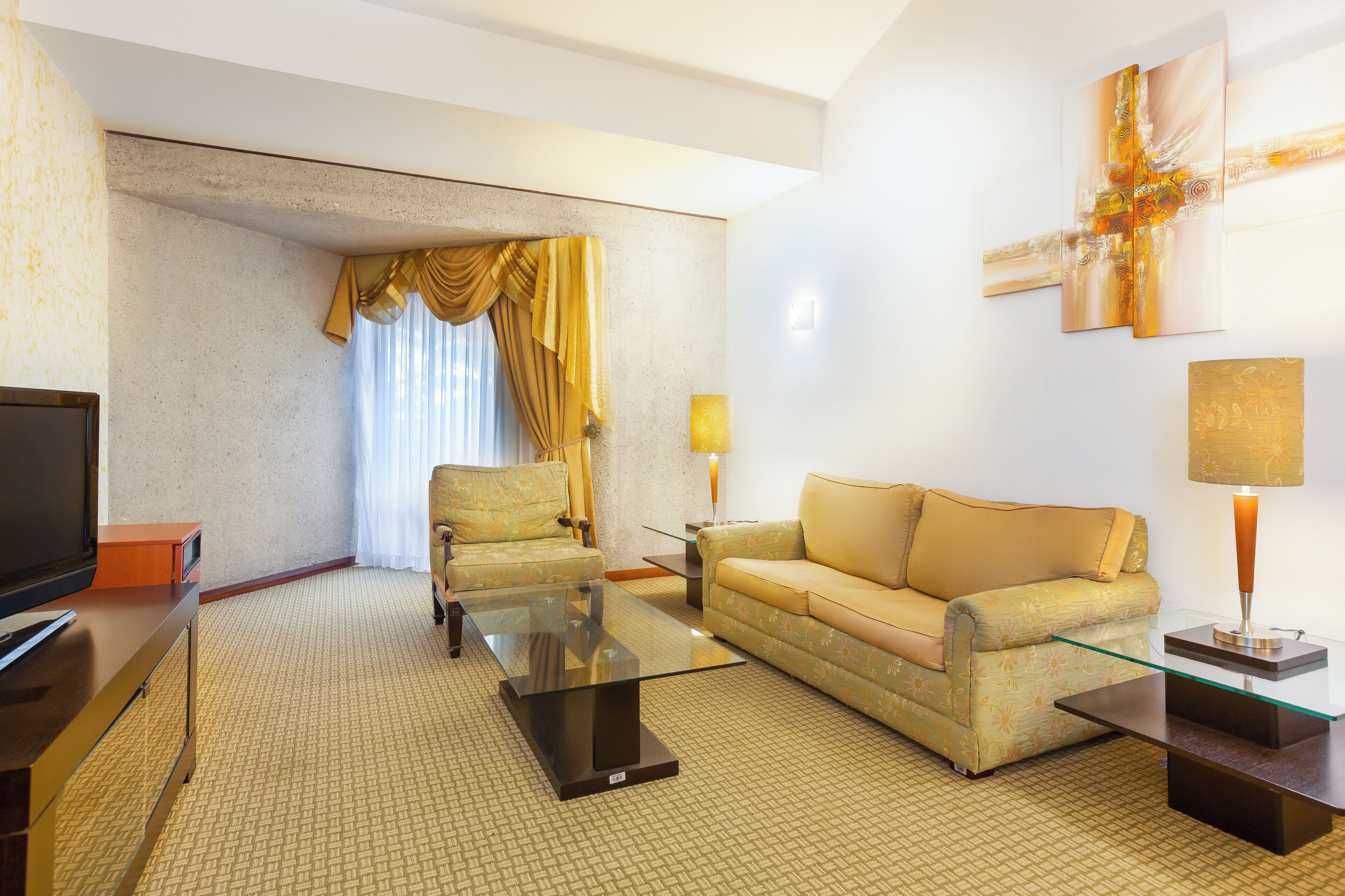 A hotel room with a sofa, armchair, glass coffee table, flat-screen TV, and a lamp.