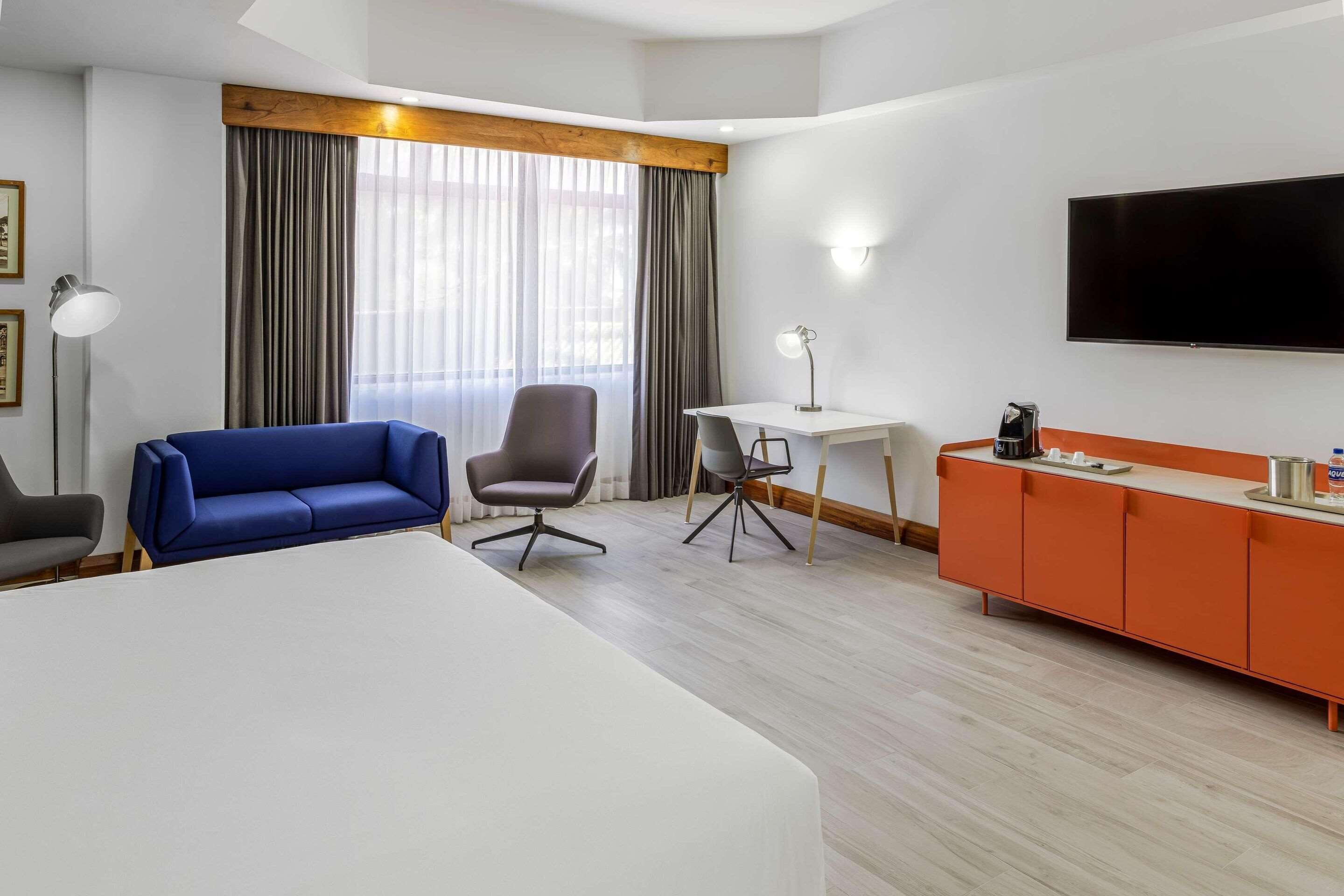 A hotel room with a bed, a blue sofa, a gray armchair, a small desk, a TV, and an orange cabinet.