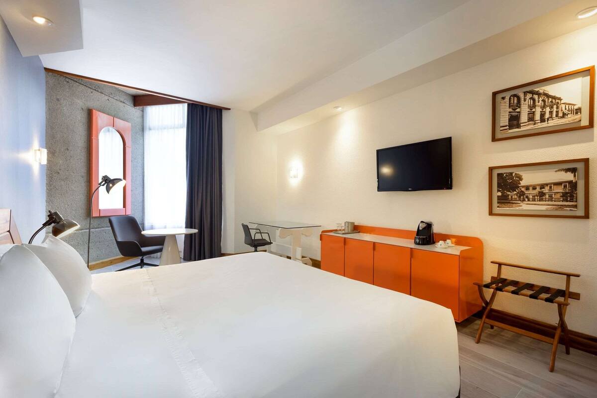 A hotel room with a large bed, a desk, a chair, an orange cabinet, a TV, and framed pictures on the wall.
