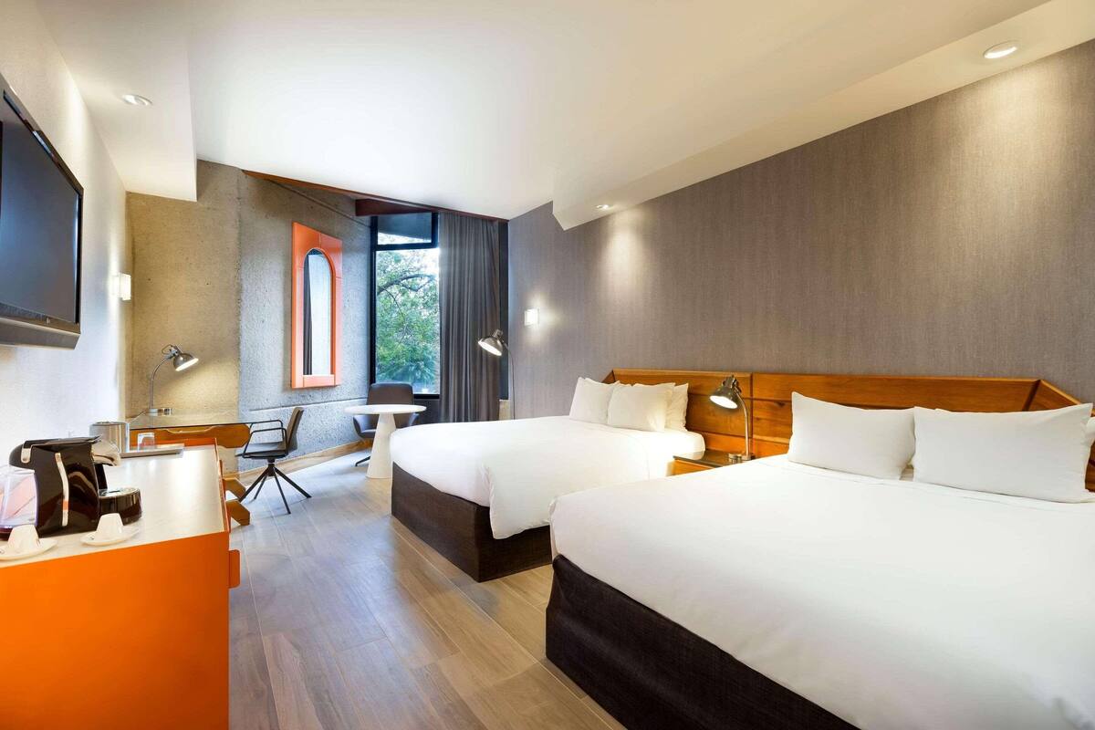 A hotel room with two beds, a desk, a chair, a TV, and a window with a view of trees.