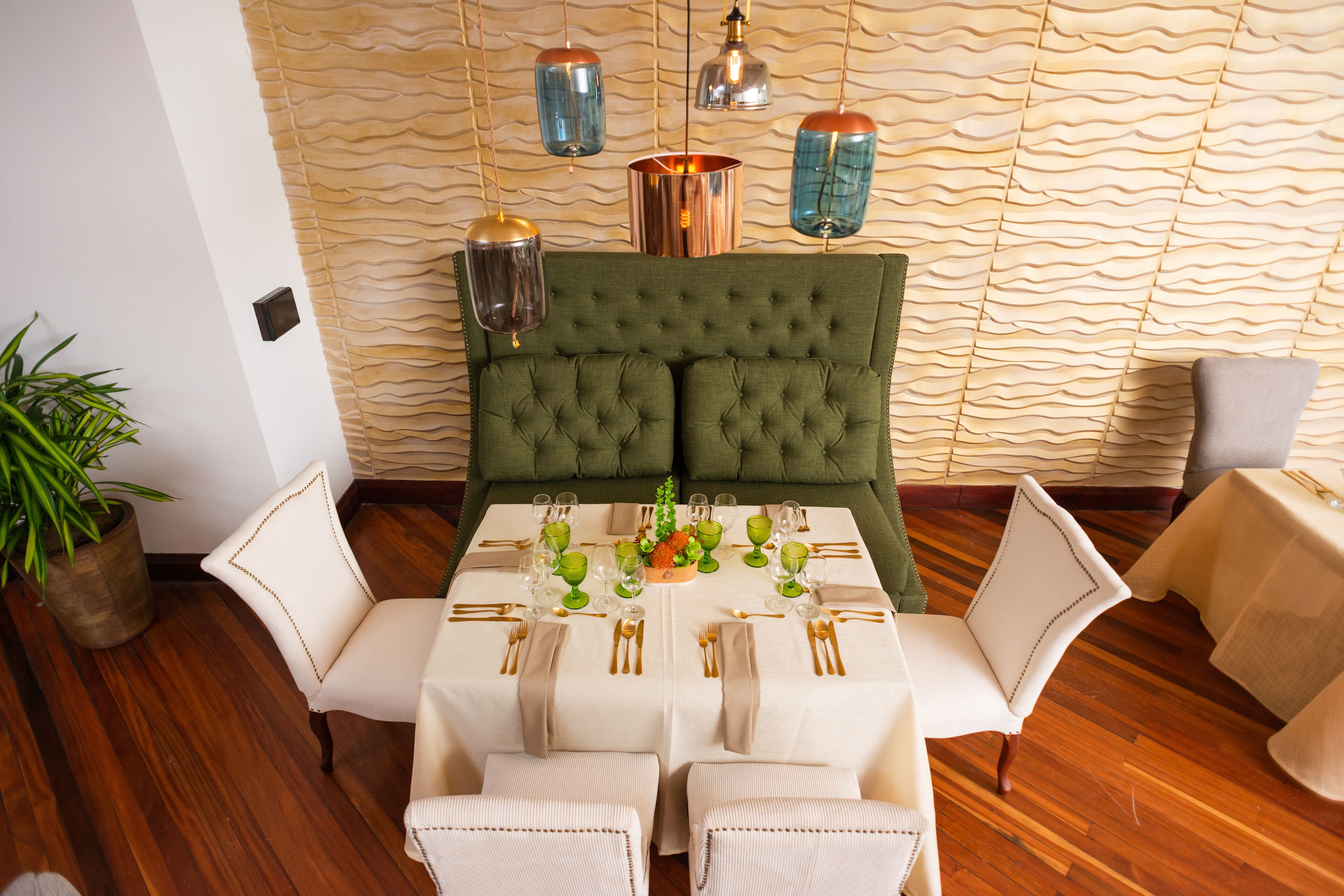 Comedor con un banco tapizado en verde, sillas blancas y una mesa preparada para cuatro, con cubiertos dorados y vasos de vidrio verdes.
