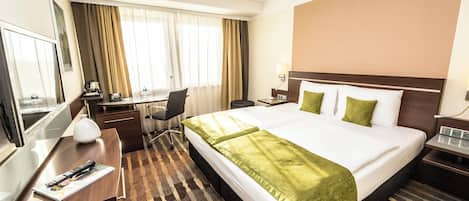 A hotel room with a large bed, a desk with a chair, a desk lamp, a TV, and a window with curtains.
