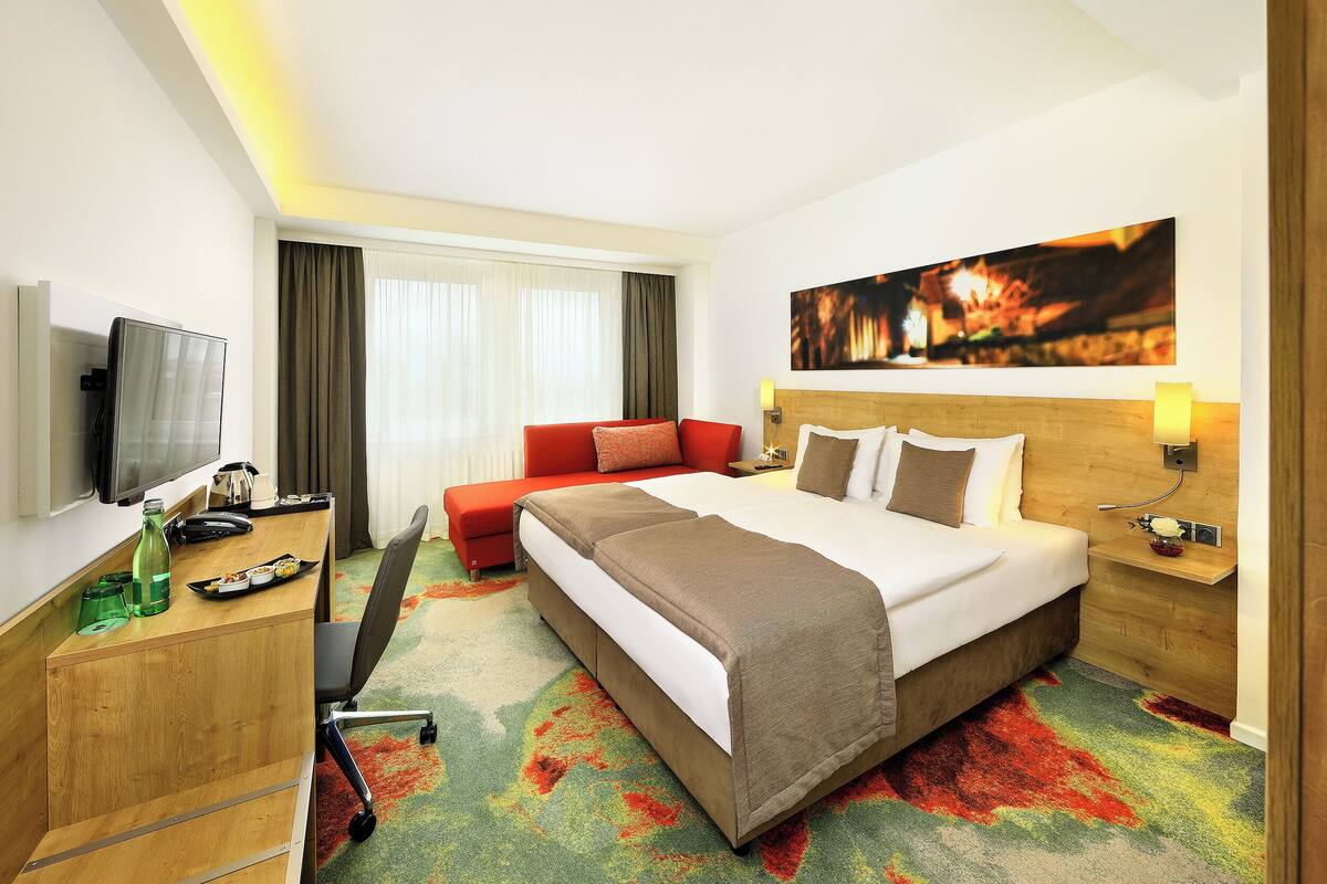 A hotel room with a bed, a desk, a television, and a large window with curtains.