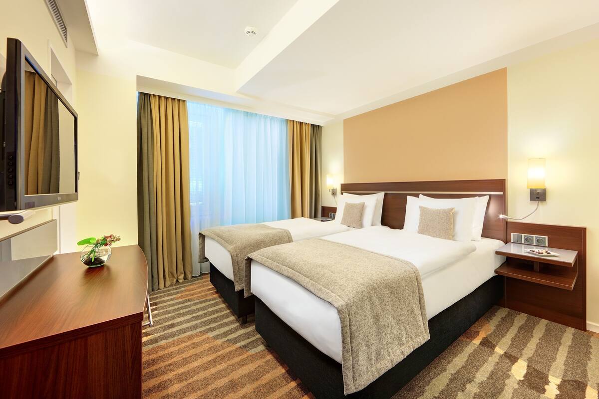 A hotel room with two beds, a desk, a TV, and a window with curtains.