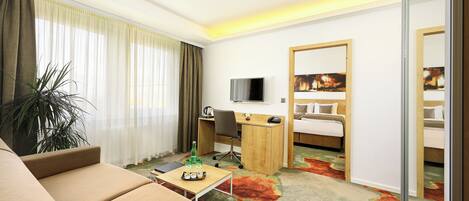 Habitación de hotel con sofá, escritorio, televisión, cama y una ventana grande con cortinas.