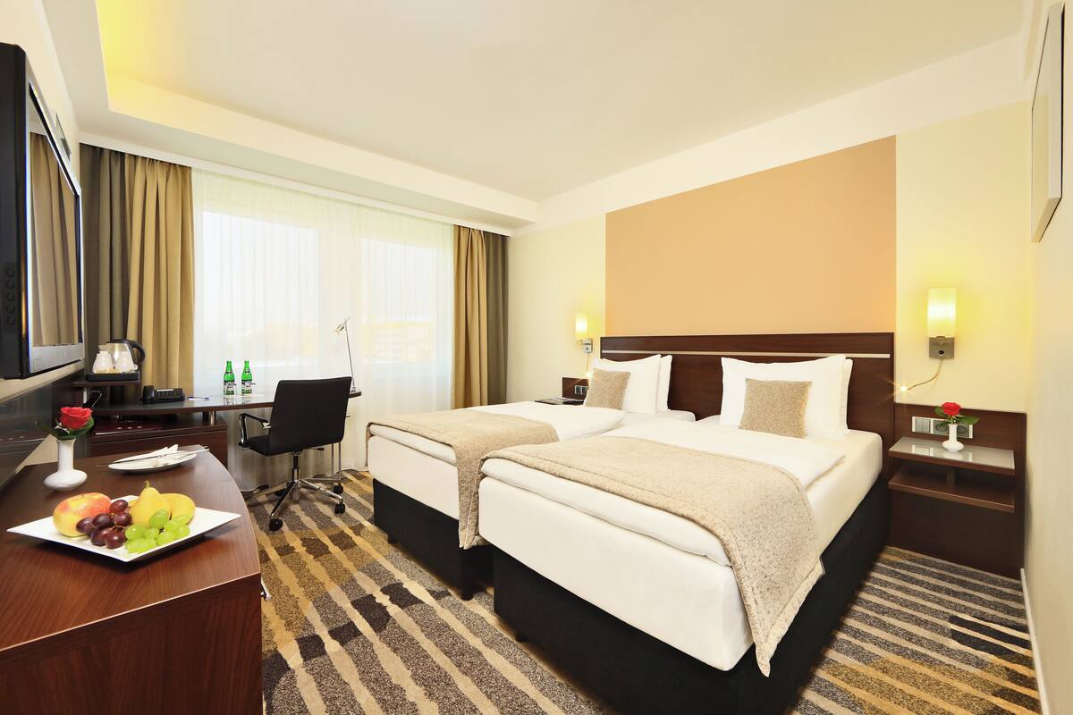 A hotel room with two beds, a desk with a computer, a chair, and a dining table with fruit.