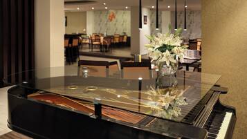 A grand piano with a glass top, a vase with white flowers, and a dining area in the background.