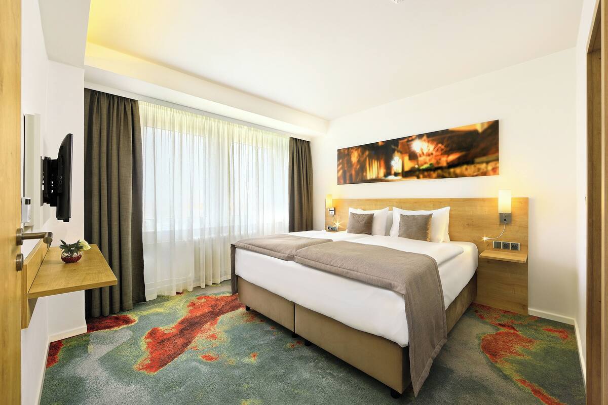 A hotel room with a bed, a bedside table, a mounted TV, and a large window with curtains.