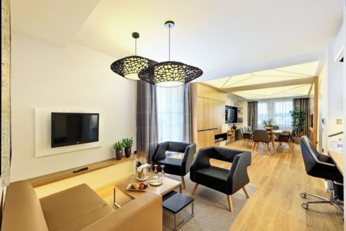 A modern hotel room with a flat-screen TV, a dining area with a table and chairs, and a kitchen area with a dining table and chairs.