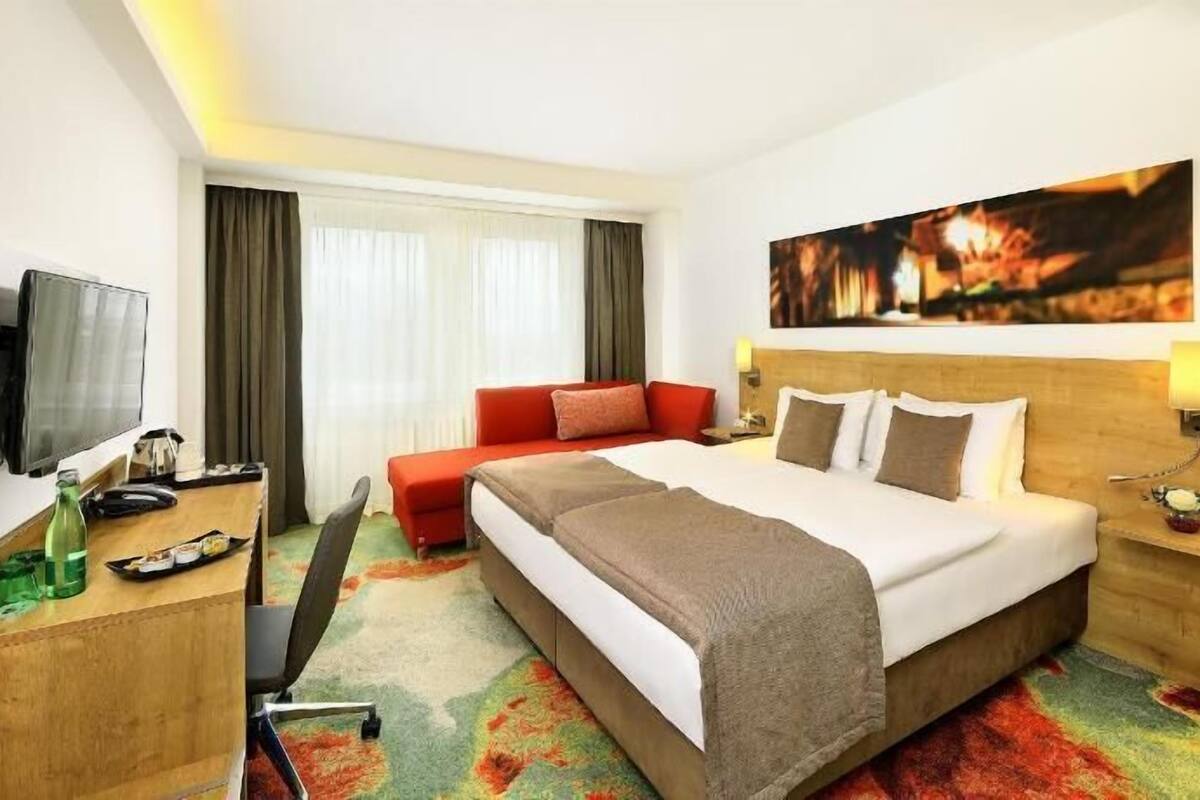 A hotel room with a bed, a desk, a chair, a television, and a painting on the wall.