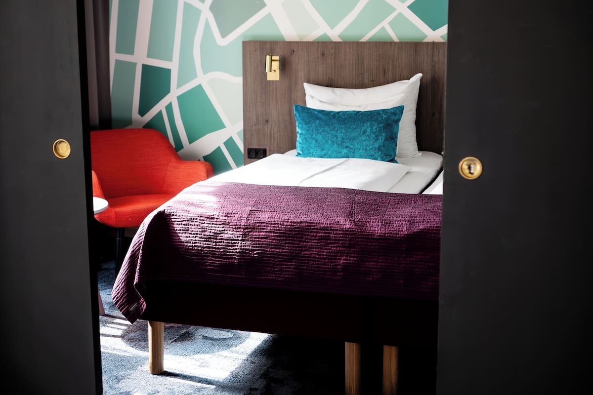 Quarto de hotel com uma cama, uma cadeira vermelha e uma parede com padrão geométrico.
