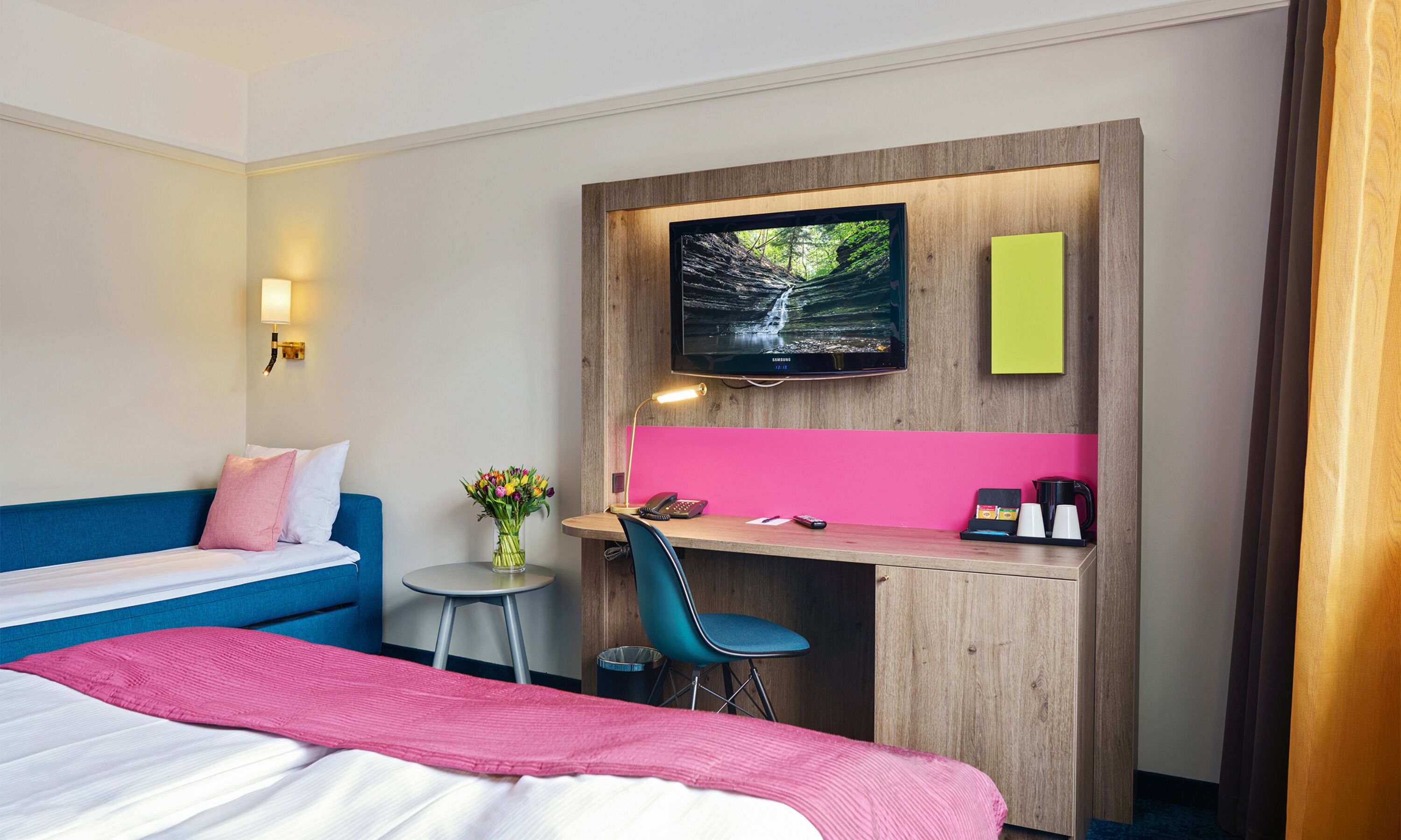 Una habitación de hotel con una cama, un escritorio con computadora, televisión y una pequeña mesa con un florero.