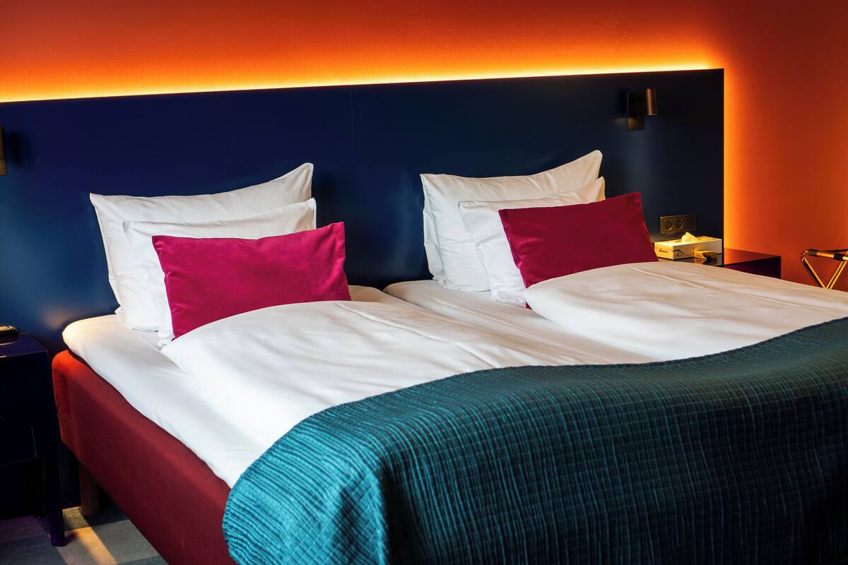 Cama de casal com roupa de cama branca e verde-água, dois travesseiros e uma mesa de cabeceira com abajur.