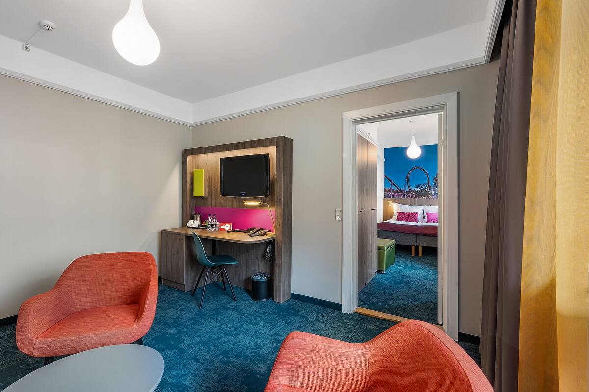 Quarto de hotel com uma escrivaninha, duas cadeiras laranja, uma cama e um espelho grande.