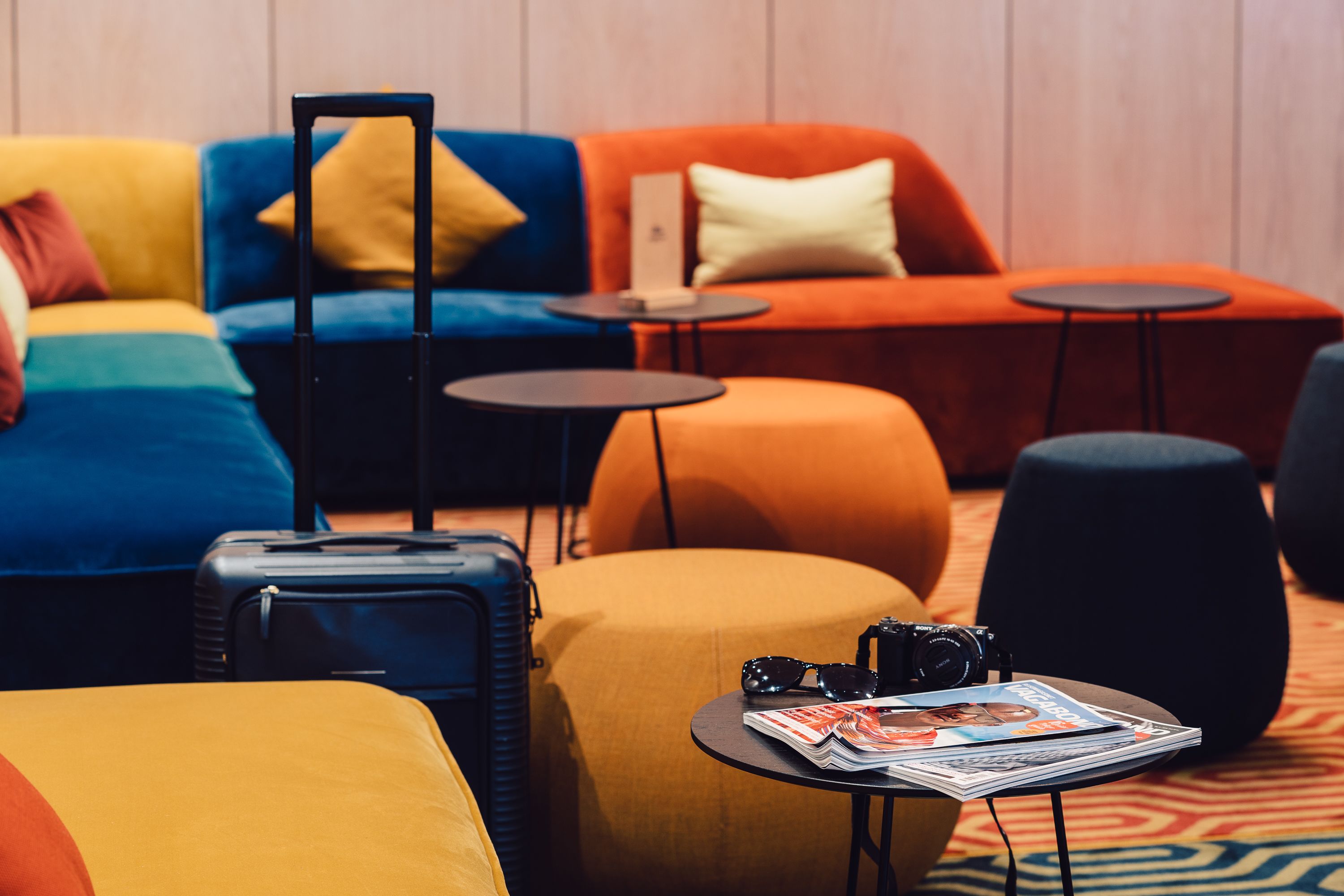 Un vestíbulo de hotel moderno con sofás coloridos, una pequeña mesa con una cámara y revistas, y una maleta.