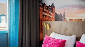 Hotellihuone, jossa on sänky, ikkuna verhoilla, seinään kiinnitetty valaisin ja näkymä kaupungin ylle.
