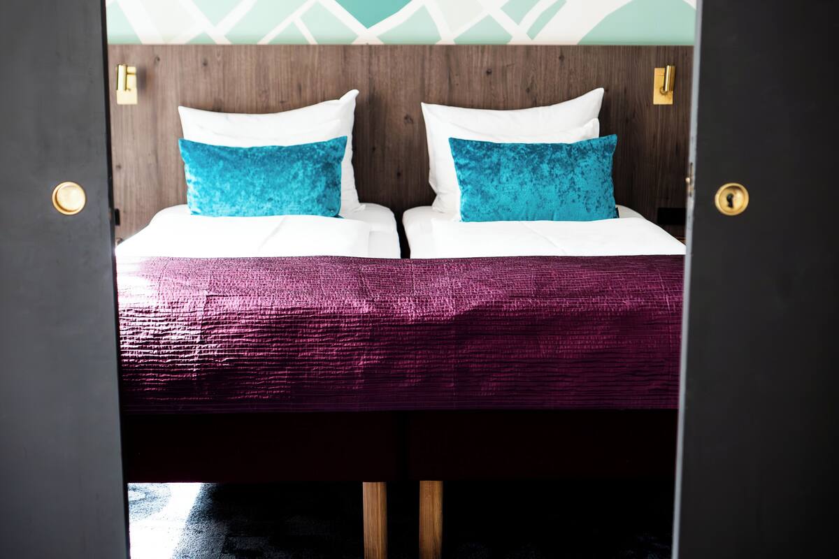 Uma cama de casal com almofadas brancas e azuis, um cobreleito roxo e uma cabeceira de madeira.