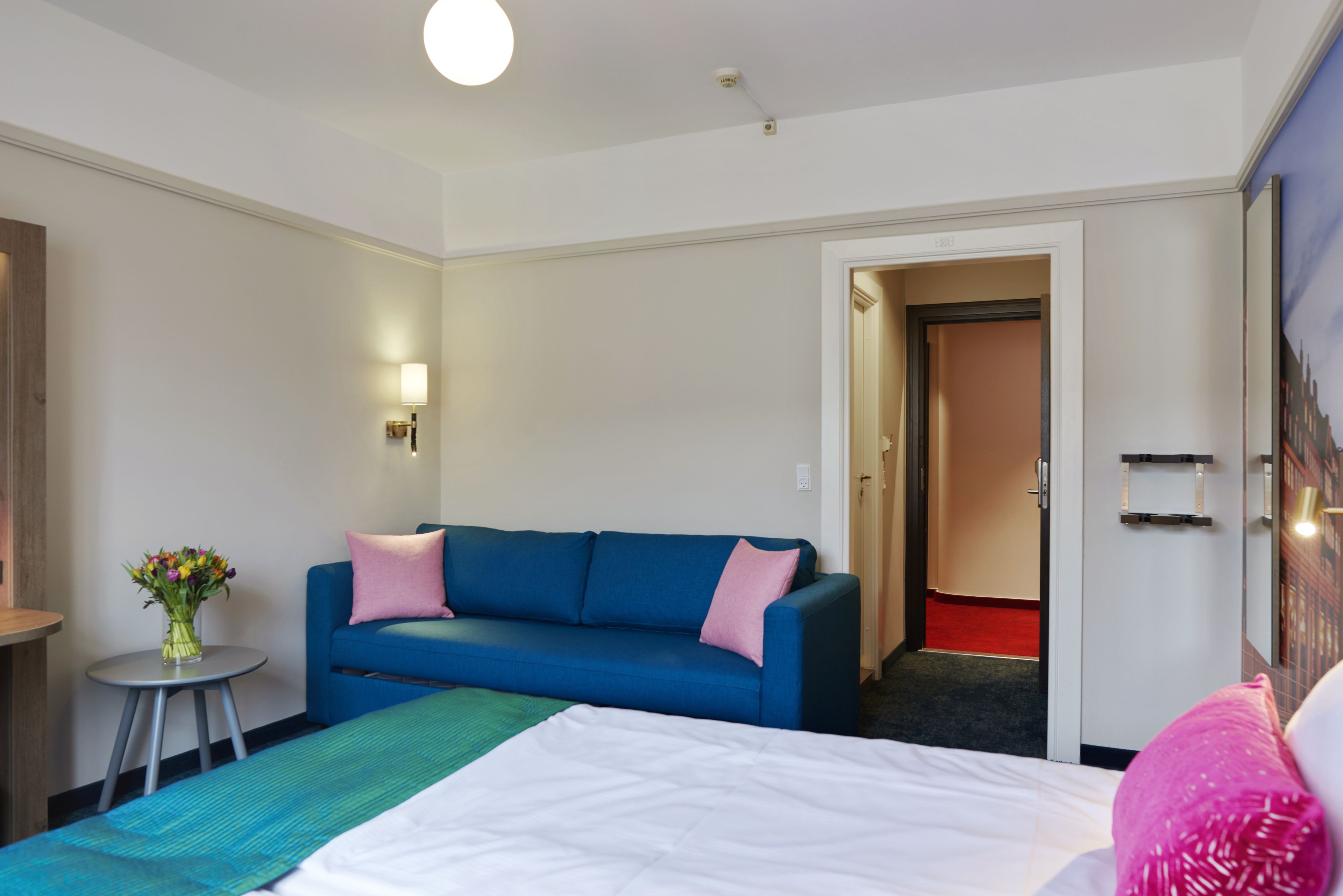 A hotel room with a bed, a blue sofa, a small table with a vase of flowers, and a door leading to another room.