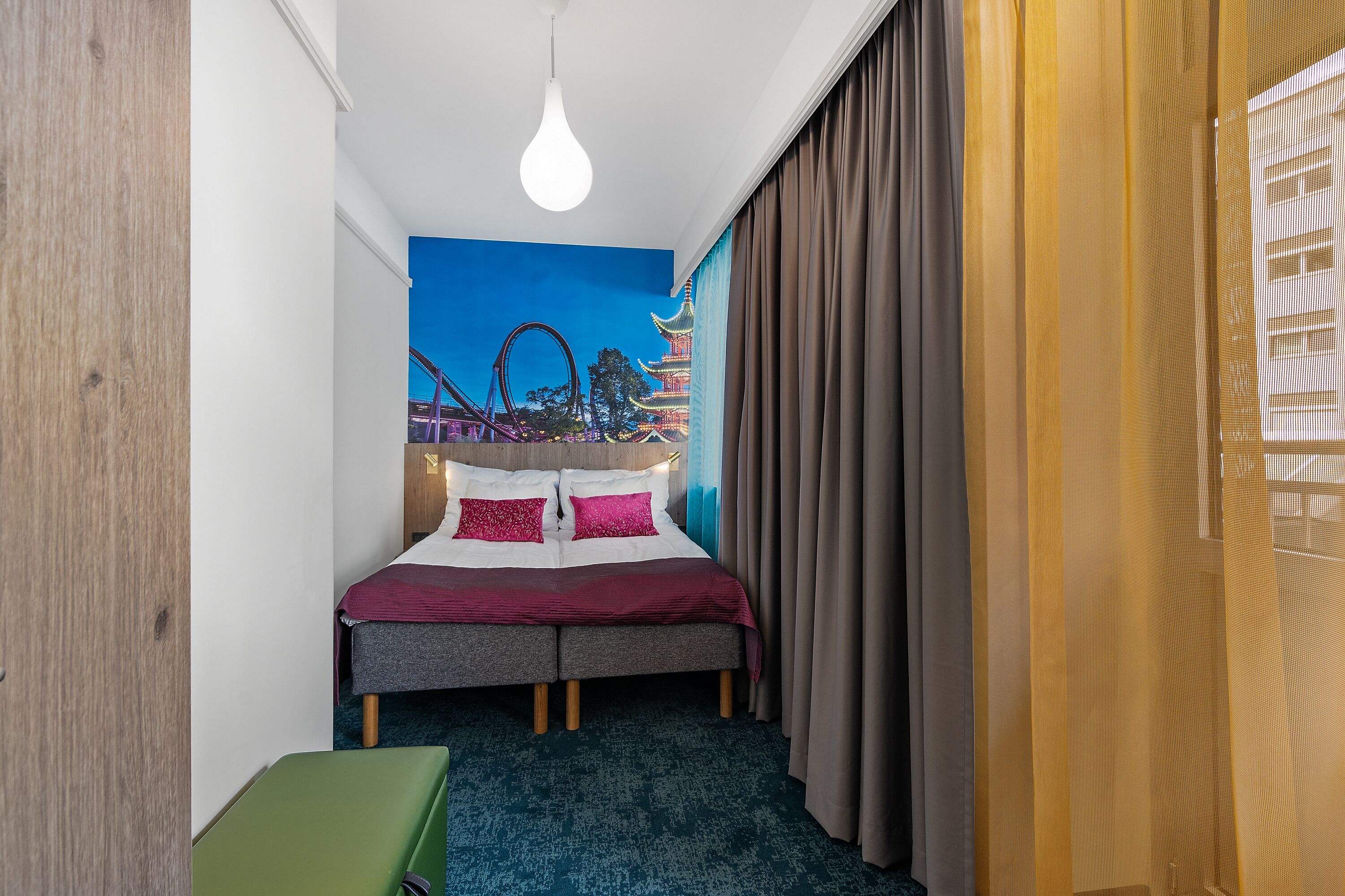 A hotel room with a bed, a green chair, a blue wall mural, and a window with curtains.