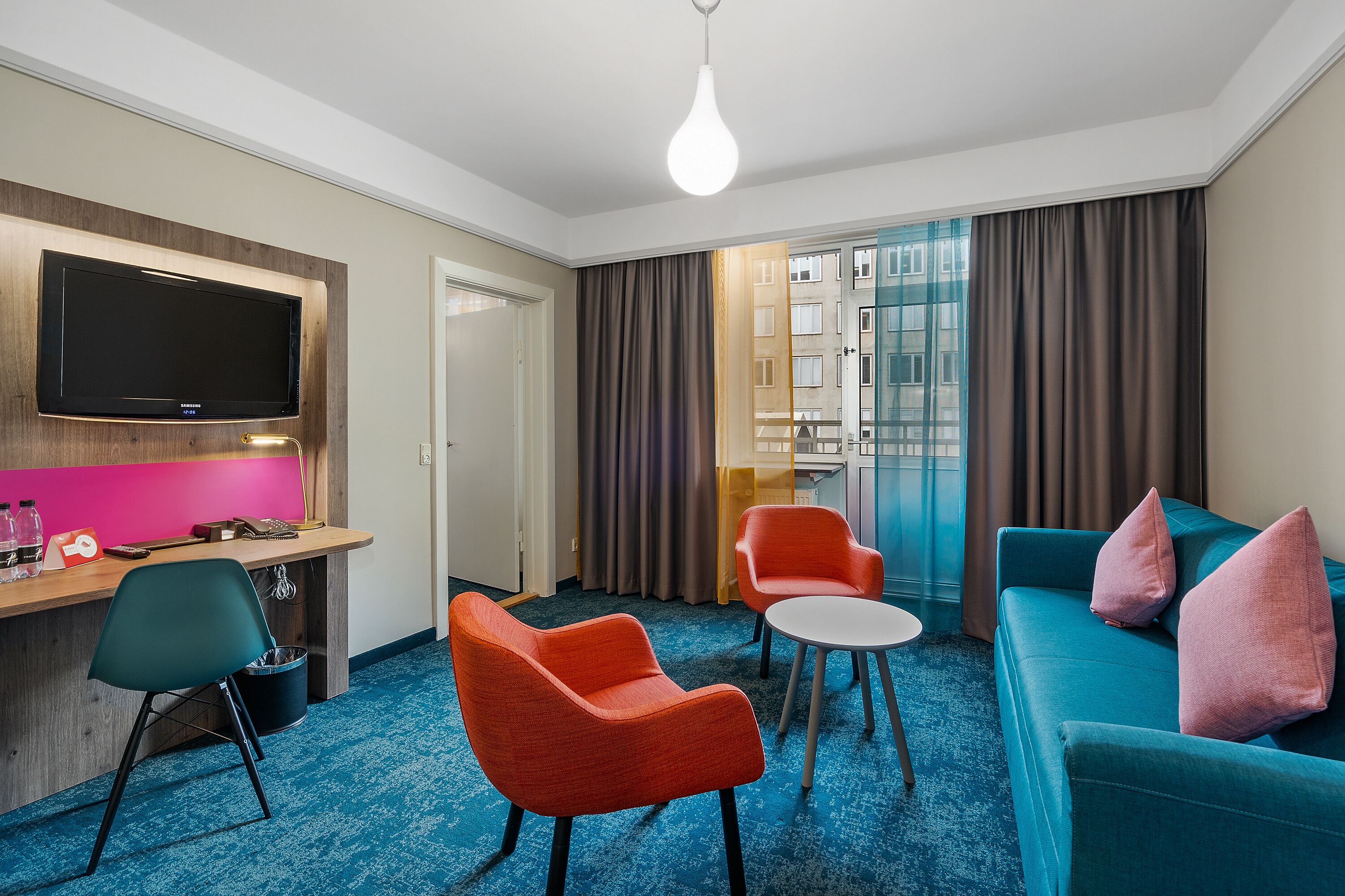 A hotel room with a TV, a desk, two orange chairs, a teal sofa, and a small round table.