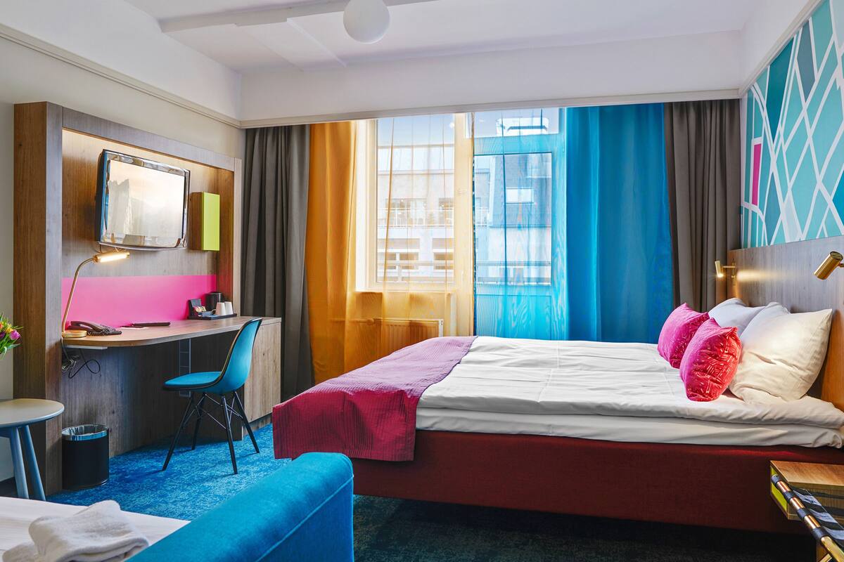 Quarto de hotel com cama, escrivaninha, cadeira, mesa pequena com um vaso de flores e vista da cidade por grandes janelas.
